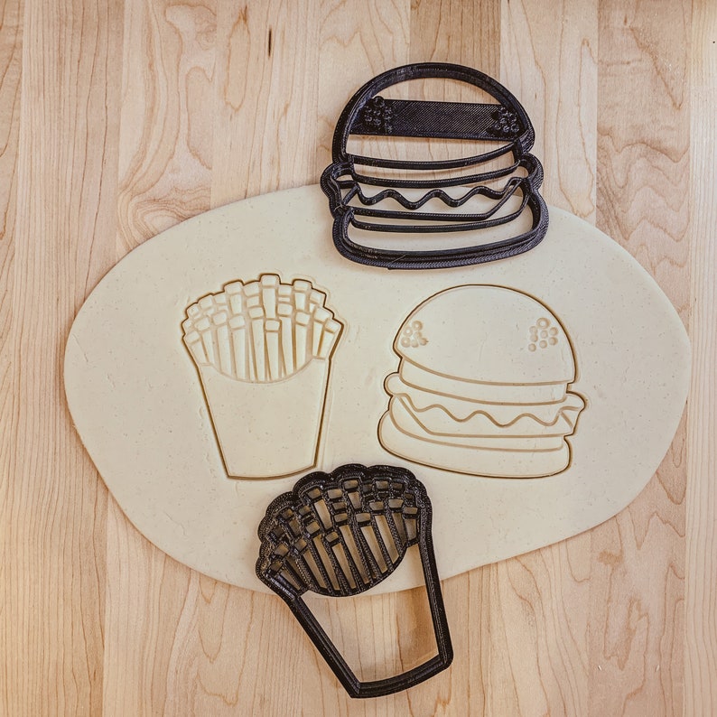 May include: Three black plastic cookie cutters shaped like a hamburger, french fries, and a burger. The cutters are on a light brown wooden surface.