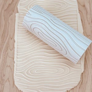 May include: A white rolling pin with a wood grain pattern is rolling over a sheet of dough, creating a wood grain texture.