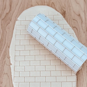 May include: A white rolling pin with a brick pattern design is rolling over a sheet of dough. The dough is also patterned with a brick design.