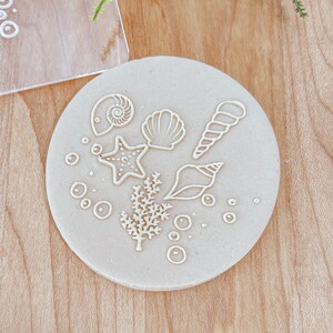 May include: A beige clay disc with white outlines of seashells, a starfish, seaweed, and bubbles.