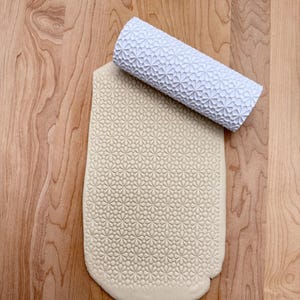 Peut inclure: Un rouleau à pâtisserie blanc à motifs repose sur une pâte à modeler, imprimée du même motif floral. Le rouleau à pâtisserie et la pâte sont posés sur une surface en bois clair. Le motif est un motif répétitif de petites fleurs stylisées.