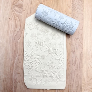 May include: A light blue rolling pin with snowflake patterns lies on a white surface embossed with the same snowflake design. The rolling pin is cylindrical and made of a smooth material. The embossed surface appears to be a sheet of dough or clay.