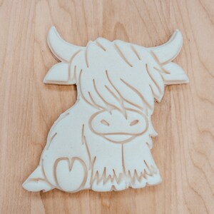 Puede incluir: Cortador de galletas de pl&aacute;stico blanco con forma de vaca de las tierras altas con un coraz&oacute;n en un lado.