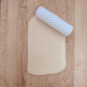 May include: A white patterned rolling pin with a diamond design lies on a sheet of dough, which also has the same pattern. The dough is a light beige colour, and the rolling pin is white. The scene is set on a light-coloured wooden surface.