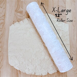 May include: A white rolling pin, 30 cm long, with a snowflake pattern, alongside a piece of dough with the same embossed design. The rolling pin is cylindrical and textured. The dough is a light beige colour.