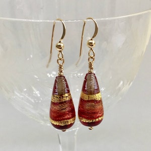 May include: A pair of gold dangle earrings with red and gold glass teardrop beads. The beads have a striped pattern with gold leaf accents.