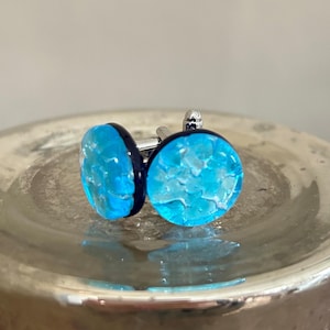 May include: A pair of cufflinks with a blue glass design. The glass has a textured, cracked ice look. The cufflinks are silver with a black border around the glass.