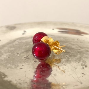 May include: A pair of gold stud earrings with red glass balls. The earrings are on a silver surface.