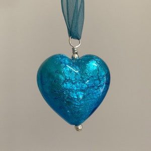 Puede incluir: Un colgante de corazón de vidrio azul turquesa con detalles plateados. El corazón tiene un acabado texturizado y craquelado.