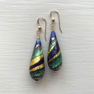 Puede incluir: Un par de pendientes en forma de lágrima con detalles de vidrio dorado y azul. Los pendientes están hechos de metal dorado y tienen una cuenta de vidrio en forma de lágrima con un patrón azul, verde y dorado.