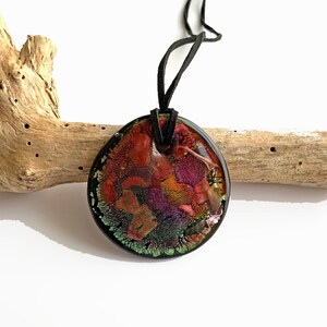 May include: A round pendant necklace with a black suede cord. The pendant is made of clear resin with embedded colourful abstract shapes and iridescent glitter.