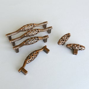 Könnte beinhalten: Satz von sechs antiken Bronze-Schrankknöpfen mit einem dekorativen Gitterdesign. Die Knöpfe sind in verschiedenen Formen und Größen, darunter zwei lange Griffe und vier kleinere Knöpfe.