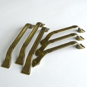 May include: Five antique brass cabinet pulls with a decorative design. The pulls are arranged in a staggered pattern on a white surface.