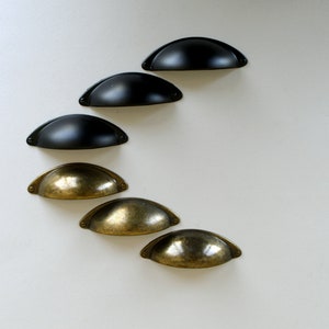 May include: Six cabinet pulls, three black and three brass, arranged in a diagonal line on a white background. The pulls are all the same shape, a half-circle with a slight curve at the top.