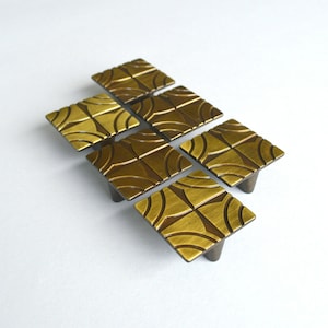 May include: Six brass cabinet knobs with a geometric pattern. The knobs are square shaped and have a brushed finish.