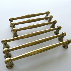 Op de afbeelding: Zes antieke messing kastgrepen. Elke greep heeft een cilindrisch middenstuk en decoratieve uiteinden. De grepen zijn gerangschikt in twee rijen van drie, wat hun ontwerp en afwerking benadrukt. Ideaal voor meubelrestauratie.