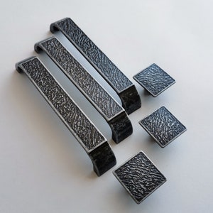 Peut inclure: Trois poignées de tiroir argentées avec une finition texturée et trois boutons carrés assortis. Les poignées sont rectangulaires et ont une extrémité incurvée. Les boutons sont carrés et ont une surface texturée.