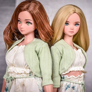 Puede incluir: Dos muñecas con el pelo largo que llevan cárdigans verdes a juego y blusas blancas con volantes. Las muñecas están de pie una al lado de la otra y mirando hacia la derecha.
