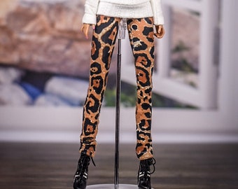 May include: A fashion doll wearing a white off-the-shoulder sweater and leopard print leggings. The doll is standing on a white display stand.