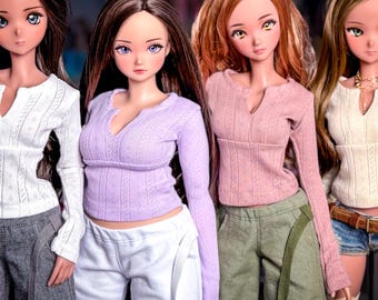 Peut inclure: Quatre poupées aux longs cheveux et aux grands yeux, portant chacune un haut à manches longues en tricot torsadé avec un col en V blanc, lavande, rose ou crème. Elles sont associées à des pantalons gris, blancs, vert olive ou un short en jean.