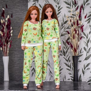 Puede incluir: Dos muñecas con pijamas a juego de color verde y blanco con estampado de tréboles, arcoíris y monedas de oro. Los pijamas tienen mangas largas y pantalones largos.