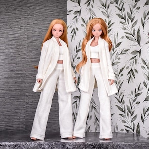 May include: Two dolls wearing white outfits with long sleeves and pants. The dolls have long, straight, light brown hair. The dolls are standing in front of a white background with a black and white floral pattern.