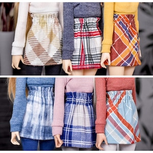 May include: A collection of doll-sized skirts in various plaid and patterned designs. The skirts feature colors like brown, red, blue, and orange. The skirts are paired with long-sleeved tops in coordinating colors, creating a stylish ensemble.