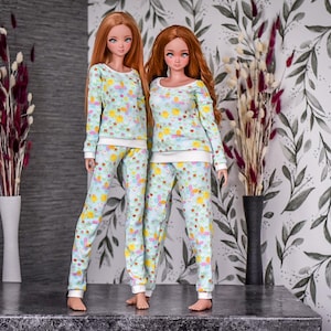 Puede incluir: Dos muñecas con pijamas a juego de color azul y blanco con estampado floral. Los pijamas tienen mangas largas y pantalones largos. Las muñecas están de pie sobre una superficie gris.