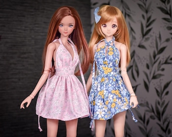 May include: Two fashion dolls wearing summer dresses. One doll has long auburn hair and a pink floral dress. The other doll has blonde hair and a blue floral dress. Both dresses have halter necklines and flared skirts.