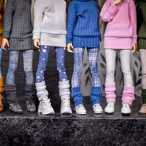 May include: A row of dolls dressed in various outfits. Each doll wears a sweater, leggings, and boots or sneakers. The sweaters are mustard yellow, gray, light gray, blue, pink, and olive green. The leggings have different patterns.