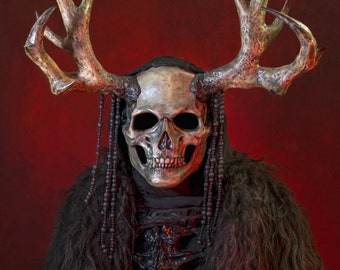 May include: A person wearing a black hooded cloak with a skull mask and antlers. The mask has a detailed bone structure and is painted in a realistic style. The person is also wearing a black fur coat and a beaded necklace.