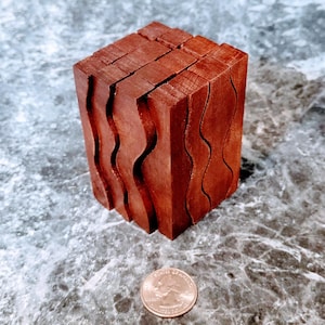 May include: A reddish-brown wooden puzzle cube with wavy, carved edges. The cube is composed of several rectangular pieces. A U.S. quarter is next to the cube for size comparison. The cube is on a gray and white marbled surface.