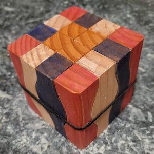Puede incluir: Un cubo de rompecabezas de madera multicolor con bloques rojos, azules y de madera natural. El cubo est&aacute; asegurado con una banda el&aacute;stica negra. El rompecabezas est&aacute; hecho de varias piezas de madera.