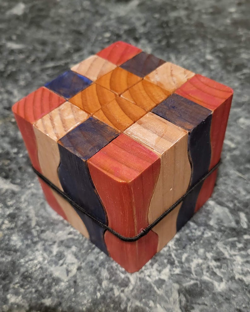 Puede incluir: Un cubo de rompecabezas de madera multicolor con bloques rojos, azules y de madera natural. El cubo est&aacute; asegurado con una banda el&aacute;stica negra. El rompecabezas est&aacute; hecho de varias piezas de madera.