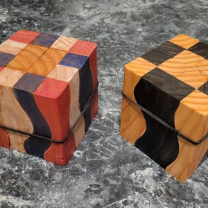 Puede incluir: Dos cajas de rompecabezas de madera. Una es un cubo con un patr&oacute;n de tablero de ajedrez en negro y madera natural. La otra tiene un dise&ntilde;o de patchwork en rojo, azul, naranja y madera natural. Ambas est&aacute;n aseguradas con una banda negra.