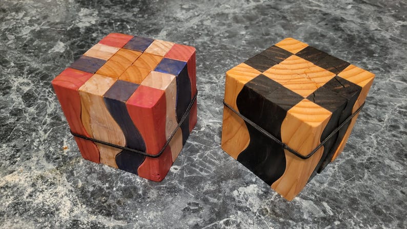 Puede incluir: Dos cajas de rompecabezas de madera. Una es un cubo con un patr&oacute;n de tablero de ajedrez en negro y madera natural. La otra tiene un dise&ntilde;o de patchwork en rojo, azul, naranja y madera natural. Ambas est&aacute;n aseguradas con una banda negra.