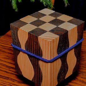 May include: A wooden puzzle box with a checkerboard pattern on top and wavy, patterned sides. The box is secured with a blue elastic band. The wood has natural and dark brown tones.