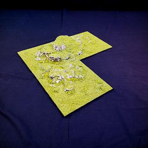 May include: A green and brown textured terrain board with a raised, rocky hill in the centre. The board is made of foam and is designed for use in tabletop wargaming.