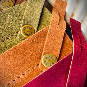 May include: Five strips of leather in different colors, each with a brass tag that reads "SUEDE CREEK". The colors are brown, green, orange, red, and burgundy.