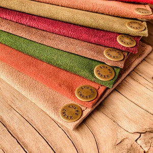 Puede incluir: Una pila de ocho carteras de cuero de gamuza en varios colores, cada una con un bot&oacute;n de lat&oacute;n que dice "Suede Creek".