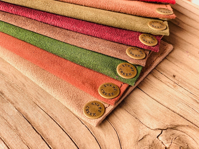Puede incluir: Una pila de ocho carteras de cuero de gamuza en varios colores, cada una con un bot&oacute;n de lat&oacute;n que dice "Suede Creek".