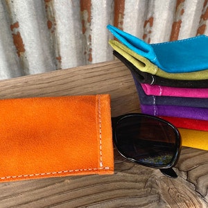 Puede incluir: Una pila de estuches para gafas de ante de colores, en varios colores, incluyendo turquesa, verde, negro, rosa, morado, rojo y amarillo. Los estuches est&aacute;n doblados y apilados sobre una superficie de madera. Un solo estuche para gafas de ante naranja est&aacute; colocado plano sobre la superficie, con un par de gafas de sol negras a su lado.