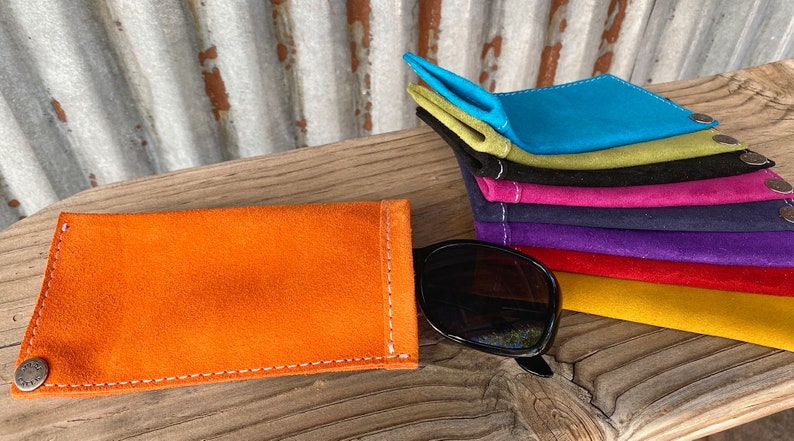Puede incluir: Una pila de estuches para gafas de ante de colores, en varios colores, incluyendo turquesa, verde, negro, rosa, morado, rojo y amarillo. Los estuches est&aacute;n doblados y apilados sobre una superficie de madera. Un solo estuche para gafas de ante naranja est&aacute; colocado plano sobre la superficie, con un par de gafas de sol negras a su lado.