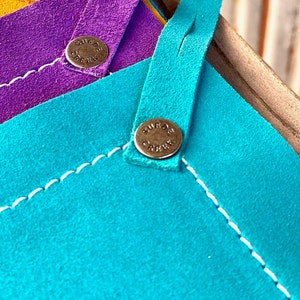 May include: Three leather squares in yellow, purple, and turquoise with white stitching around the edges. Each square has a brass button with the words "Surf City" engraved on it.