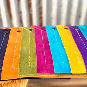 May include: A set of ten colorful leather placemats with white stitching and a button detail. The placemats are arranged in a rainbow pattern, with black, red, navy blue, orange, green, magenta, light blue, yellow, purple, and turquoise.