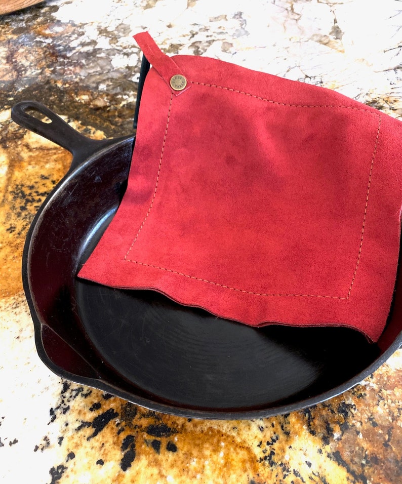 May include: A red leather pot holder with a stitched edge and a brass button. The pot holder is sitting on top of a black cast iron skillet.