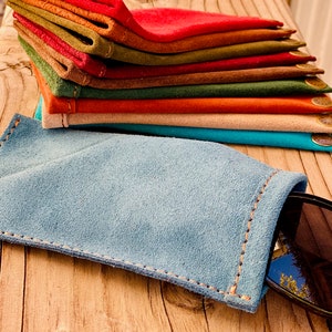 Puede incluir: Una pila de estuches para gafas de ante de varios colores, en tonos de rojo, naranja, marr&oacute;n, verde y azul. Los estuches est&aacute;n doblados y apilados sobre una superficie de madera. Un estuche para gafas de ante azul est&aacute; sobre la superficie con unas gafas de sol dentro.