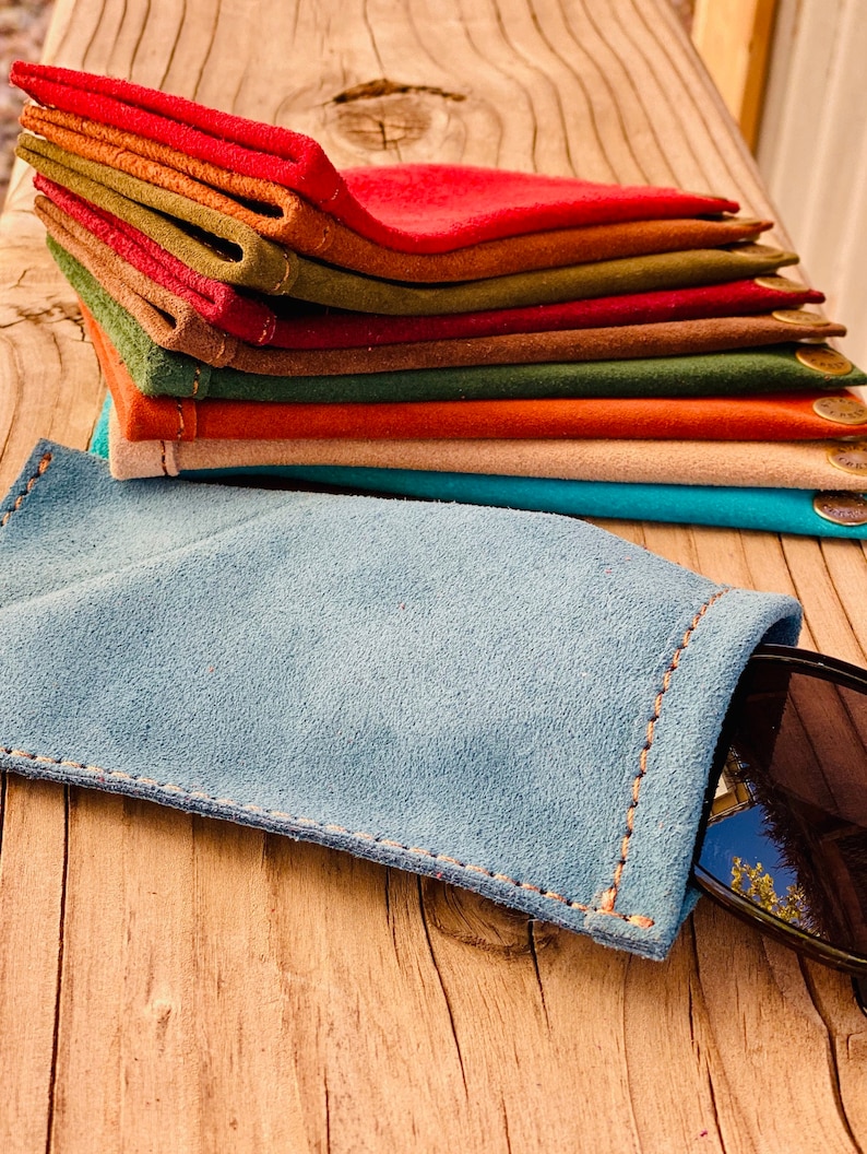 Puede incluir: Una pila de estuches para gafas de ante de varios colores, en tonos de rojo, naranja, marr&oacute;n, verde y azul. Los estuches est&aacute;n doblados y apilados sobre una superficie de madera. Un estuche para gafas de ante azul est&aacute; sobre la superficie con unas gafas de sol dentro.