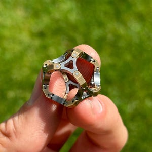 May include: A multi-toned metal ring with a complex geometric design. The ring features silver, gold, and bronze-colored segments, creating a layered and intricate appearance. The ring is held against a blurred green background.
