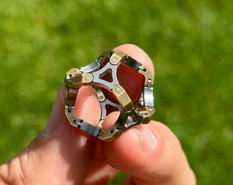 May include: A multi-toned metal ring with a complex geometric design. The ring features silver, gold, and bronze-colored segments, creating a layered and intricate appearance. The ring is held against a blurred green background.